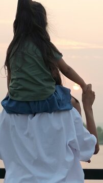 Back view of a caring father carrying his young daughter on his shoulders, watching the sunset together near a lake, symbolizing love, family bonding, and cherished childhood memories
