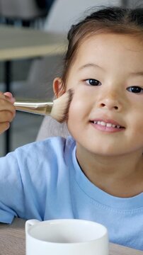 Adorable little asian girl with dark hair styled in buns sitting at a table and enthusiastically applying cosmetic powder to her face with a large brush, exploring her mother's makeup