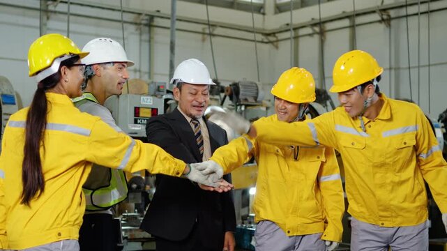 Adult diverse factory workers and a manager in hard hats stack hands together in a huddle, cheering and clapping in a manufacturing plant, celebrating successful teamwork and industrial business goals