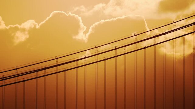 Sunset over a suspension bridge with cables and clouds illuminated by golden light, showcasing the intricate design and structure against a vibrant sky