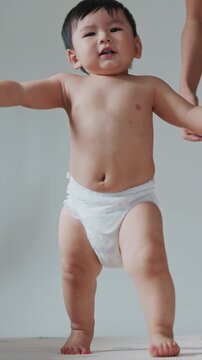 Happy Asian baby boy in diaper learning to walk with arms raised for balance.