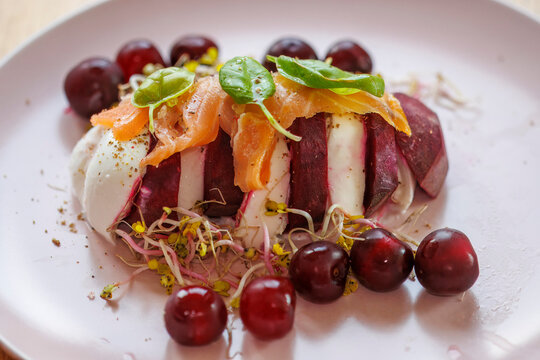 Pieces of burrata cheese interspersed with pieces of red beetroot, sprinkled on top with sprouts, basil and sliced ​​salmon on a plate with sprigs, a light summer dish.