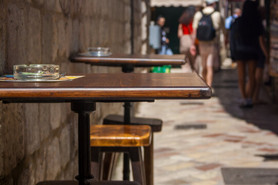 Empty wooden tables with ashtrays in a narrow street. People walking in the background. Bright sunlight illuminates the scene.