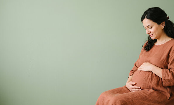 Pregnant woman in terracotta dress seated cradling belly green wall