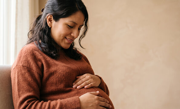 Pregnant Latina woman in rust sweater cradling belly by window