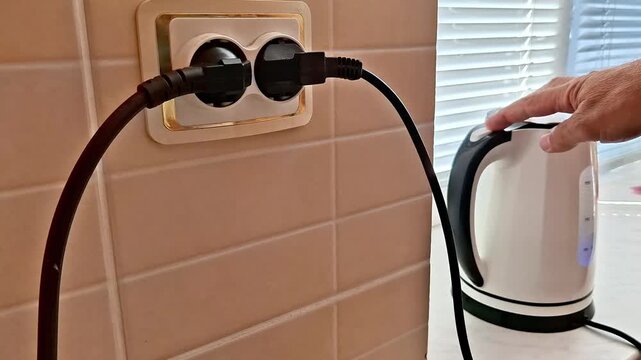 Kitchen wall outlet with two power cords plugged in as hand holds electric kettle nearby, highlighting household electricity use, appliance safety, and everyday utility