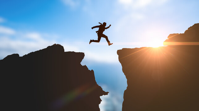 Businessman silhouette jumping across mountain gap during bright sunrise representing corporate risk extreme challenge ultimate success overcoming business obstacles achieving goals