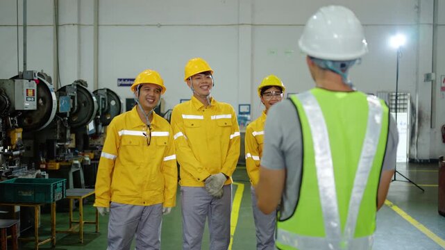 Caucasian industrial supervisor in a safety vest instructs a team of smiling factory workers wearing yellow hard hats during a morning production meeting on the manufacturing plant floor. Safety first