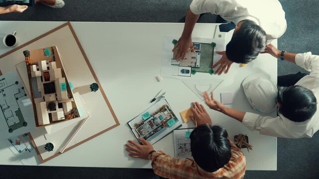 Group of skilled designer looking at model while discussing about building. Top view of architect engineer explain idea while talking and writing blue print scattering at meeting table. Alimentation.