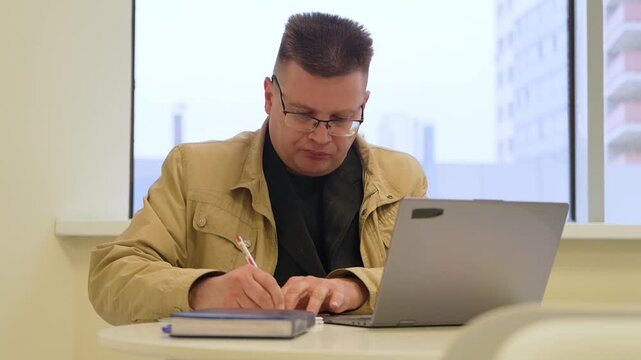 Businessman writing notes while working in a cafe with an open laptop. Remote work, flexible workplace, business planning, productivity, and modern professional lifestyle.