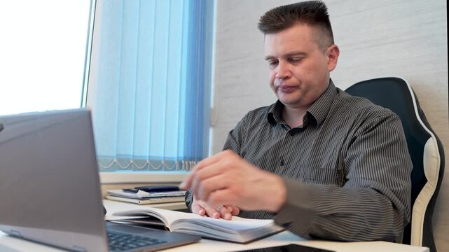 Businessman sitting at computer flipping through planner with tasks and organizing schedule, multitasking professional.