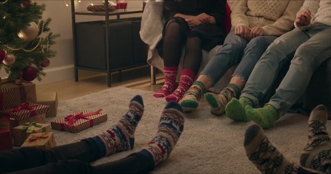 People wearing colorful knitted socks playfully moving to music around the Christmas tree with wrapped gifts in a warm home interior.