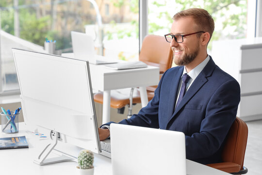 Smiling businessman in a navy suit and glasses working on a desktop computer in a modern office setting, He is focused on the screen, with a laptop and other office supplies visible on the white desk