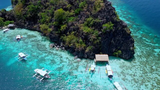 Retreating flight revealing outrigger boats anchored over shallow coral reef along the base of a dramatic karst island with turquoise waters. Coron, Palawan, Philippines
