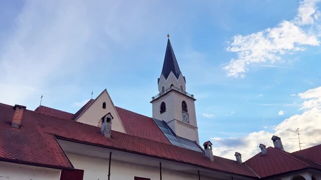Church of St. Cantianus in Kranj old town, Slovenia, with red tiled rooftops, clock tower, Gothic spire, and bright blue evening sky.