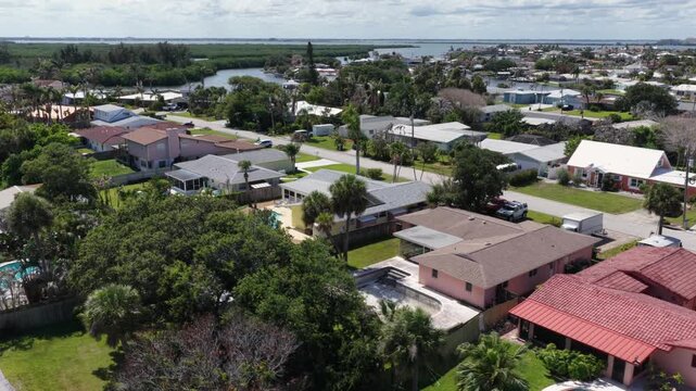 Red tile roof estate features kidney-shaped pool in backyard, palm-lined neighborhood street stretching toward mangrove canal plus bay waters in Cocoa Beach, Florida. Aerial drone wide shot