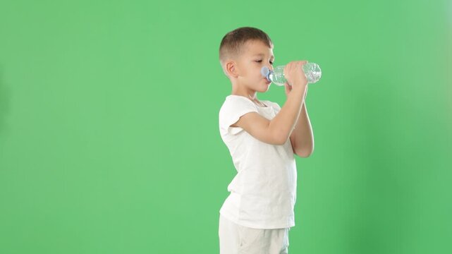 Boy in white clothes drinks water from a plastic bottle on a green background. High quality 4k footage