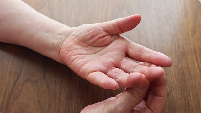 An elderly woman touching her hand on a wooden table, concerned about the pain.