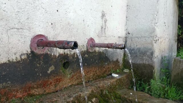 Natural spring water flowing from old metal pipes into stone trough, handheld shot