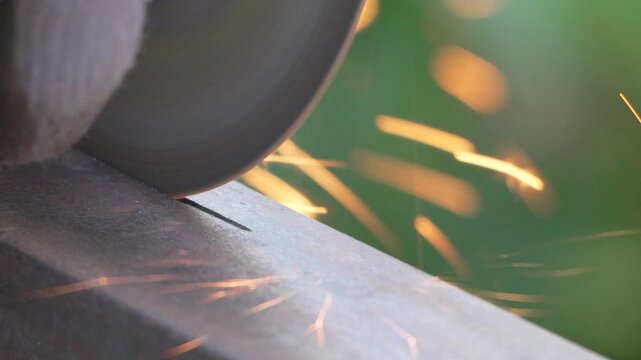Industrial angle grinder cuts through thick iron rail outdoors as friction creates intense hot luminosity. Extreme close-up shot.