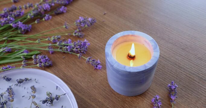 Scented lavender candle burning on wooden table background. Purple flower petals lying near blue concrete candle holder. Relaxing aromatherapy spa concept video with calming ambient mood.