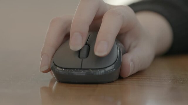 A close-up of a woman's hand clicking a dark wireless mouse on a wooden desk surface in a warm workspace.