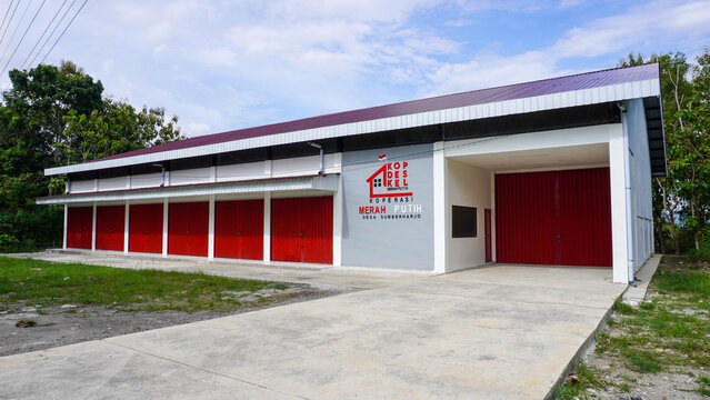 Building facade of cooperative 'koperasi merah putih' in indonesia, featuring red roller doors and white walls with large signage and an indonesian flag.