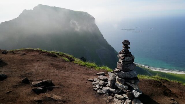 Stone cairn stands on a high dirt lookout above a sheer coastal slope with beach and open sea fading into mist in Lofoten.