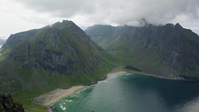 Kvalvika Beach curves below steep green mountain walls and dark bay water viewed from the Ryten ridgeline on an overcast day.
