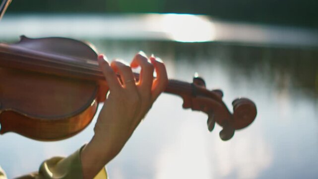 Close up of musician playing violin outdoors near water, peaceful hand movements on strings creating gentle melody in warm natural sunset lighting