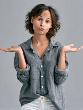 Young woman shrugging hands, casual gray shirt, white pants, neutral background, uncertain expression, short brown hair, studio portrait