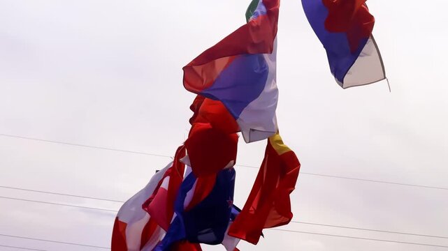 Colorful international flags waving in the wind, with visible French, Armenian, Ethiopian and Latin American flag colors against a pale sky.