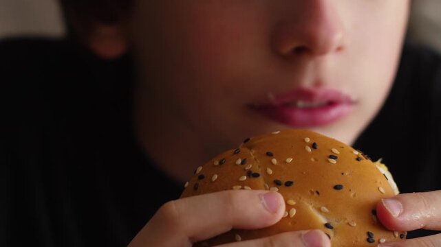 A close-up of a boy licking his fingers and appetizingly biting a juicy burger with a sesame bun. The child chews fast food with obvious enjoyment during a satisfying snack.