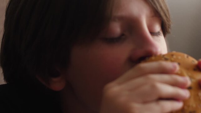 A close-up of a boy appetizingly biting and eating a juicy hamburger with a sesame bun. Closing his eyes with pleasure, the child texturfully chews fast food during lunch.
