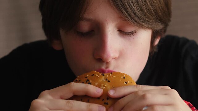 Close-up of a young boy taking a big bite of a juicy burger with his eyes closed. The child thoroughly enjoys chewing delicious fast food during a satisfying meal break.