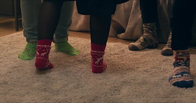 People in cozy knitted socks gather around a decorated Christmas tree with wrapped gifts in a warm living room.