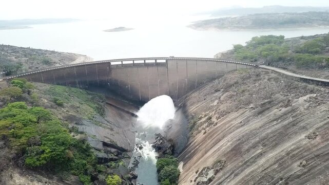 Aerial view of a large concrete arch dam releasing water through the spillway into the canyon below