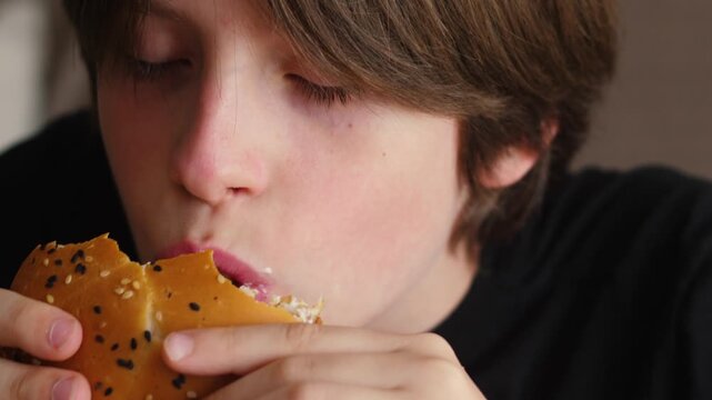 Close-up of a young boy opening his mouth wide to take a big bite of a juicy burger. The child appetizingly chews delicious fast food during a satisfying meal break