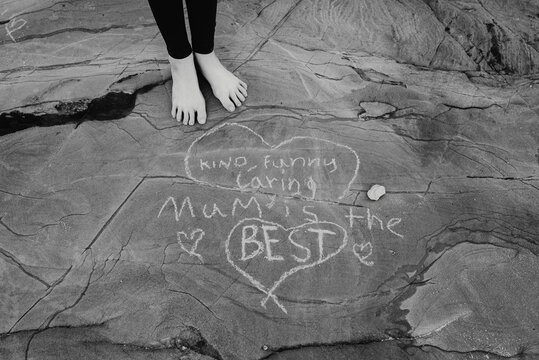 Mum is the best (kind, funny, caring). Young girl's feet standing next to a message written to her mum on rocks at the beach; Australia