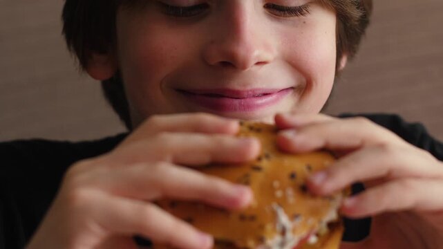 Close-up of a happy young boy holding a delicious hamburger with sesame seeds. Smiling kid enjoying fast food and about to take a bite of a juicy burger in a cafe.