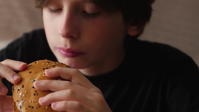 Close-up of a young boy holding a delicious hamburger with sesame seeds after taking a bite. The kid is enjoying his juicy fast food and looking down at the burger in anticipation of the next bite