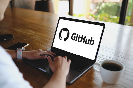 Bangkok, Thailand. July 12, 2026; An over the shoulder view of a person using a MacBook Pro laptop displaying the GitHub logo on screen at a wooden table next to a cup of coffee.