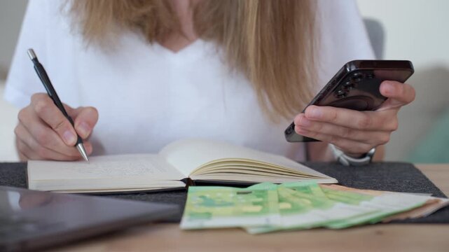 Woman managing home budget writing notes with phone and euro cash nearby. Concept of household expenses bills and personal finance.