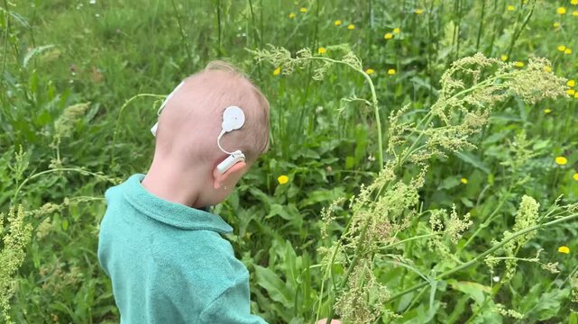 Happy little boy with cochlear implant walking through green summer grass, enjoying nature and freedom. Childhood, hearing technology, inclusion and healthy outdoor lifestyle concept.