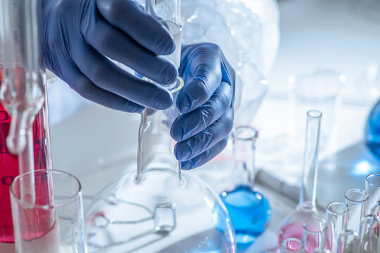 Chemistry laboratory scene with colorful chemical solutions in glass flasks and cylinders, scientist in gloves writing results, analytical experiment workspace.
