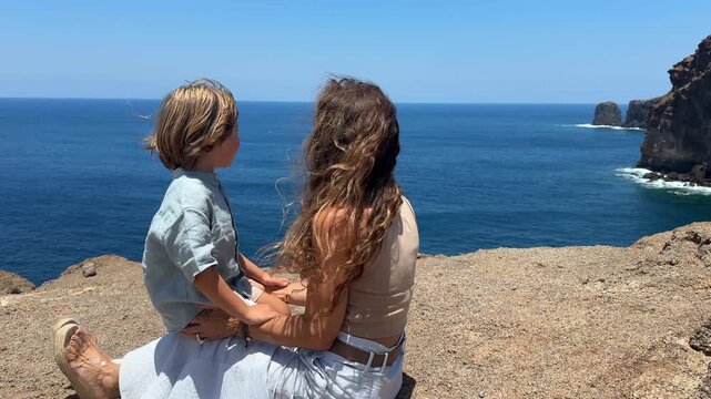Mother and son sitting on a cliff in Gran Canaria enjoying the coastal view during family holidays on a windy sunny day