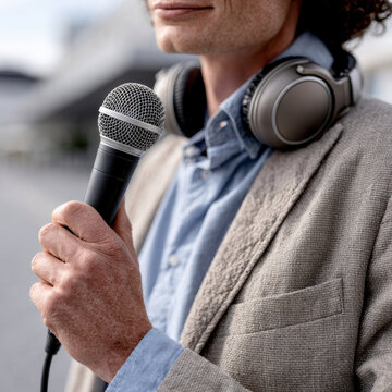 Man holding microphone wearing headphones, outdoor interview, close up, professional journalist, casual blazer, blue shirt, confident expression