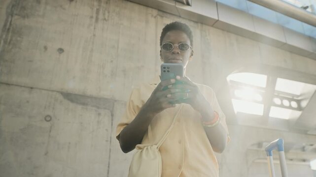 African American young adult woman wearing sunglasses standing near concrete wall, using smartphone next to blue suitcase. Video is perfect for travel apps, mobile booking, and tourism marketing
