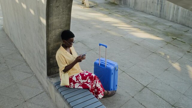 Young adult African American woman sitting on concrete bench next to blue suitcase, typing on smartphone. Clip is ideal for commercials promoting travel apps, mobile booking, or tourism services