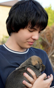 Teenage boy with black hair looking down with a gentle smile at a small newborn puppy sleeping peacefully in his arms outdoors